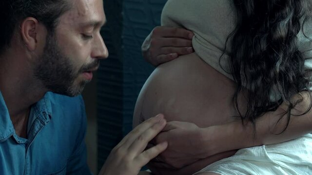 Caucasian man kissing and listening his newborn baby through tummy of pregnant wife in bedroom. Couple family spending time together at home. Family relationship and maternity healthcare concept.