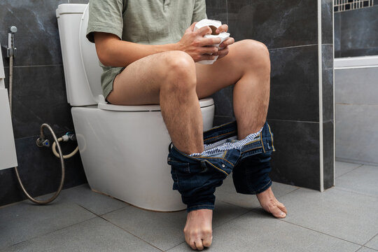 man with stomach issues sitting in toilet bowl and holding tissue roll in bathroom