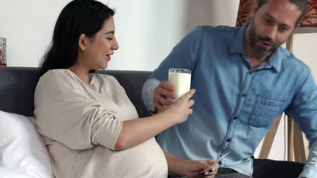 4K Caucasian man serving glass of milk to pregnant wife on the bed while looking at newborn baby ultrasound scan picture together with smiling. Happy family and maternity expecting healthcare concept