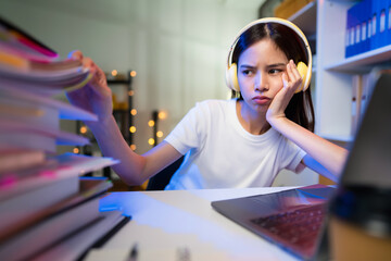 Stressful  young Asian woman wearing headset and sitting on the chair with a pile of papers document on the table and looking on the computer screen on night.