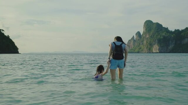 Mother lovingly holding hand with her young daughter on outdoor beach on long summer vacation. With beautiful views by the front to see the mountains, emerald seawater, sand, and blue sky.