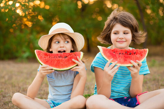 Two preschool smiling child sister sit with crossed legs on grass and eating watermelon at summer park with sunset light