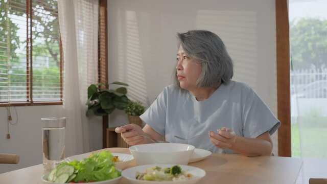 Asian senior woman feeling lonely, eating food alone in the kitchen.
