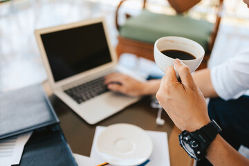 businessman holding coffee cup in hands and computer on table in coffee shop. Young freelancer drink coffee and working.
