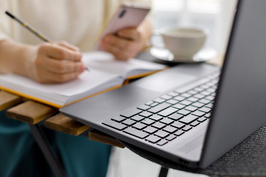 business woman works at laptop, holds smartphone in her hand and makes entries in diary. Business planning and tasks for day or week. Female in business. Keyboard laptop