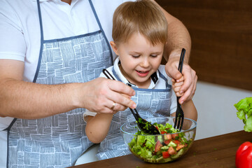 The blonde-haired son and father prepare a salad of colorful, fresh vegetables together in the kitchen. They both wore blue aprons.