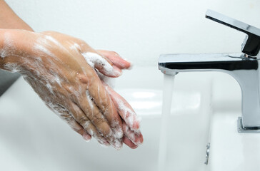 Fototapeta premium Hands of woman wash their hands in a sink with foam to wash the skin and water flows through the hands. Concept of health, cleaning and preventing germs and coronavirus from contacting hands