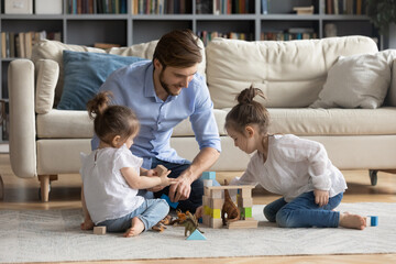 Fototapeta premium Loving Caucasian father relax in living room at home play with small daughters with blocks toys. Happy dad engaged in playful funny game activity with two little girls children. Family time concept.