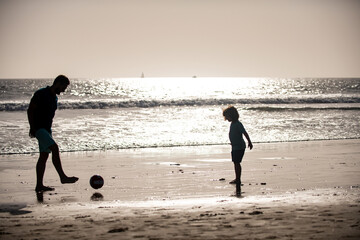 Father and son play soccer or football on the beach on summer family holidays. Dad and child enjoying outdoor silhouette on sunset.
