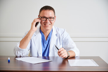 Joyful man surgeon in glasses smiling while sitting at the table with papers. Male physician holding pen and talking on cellphone in clinic. Concept of medicine, communication and healthcare workers.