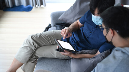 Two young man in protective mask using digital tablet while sitting together on sofa.