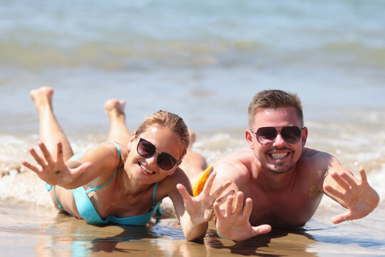Joyful man and woman in sunglasses lie on wet sand near sea