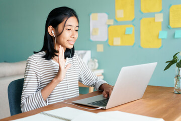 Happy Asian girl waving in greeting online tutor on tablet with headphones 