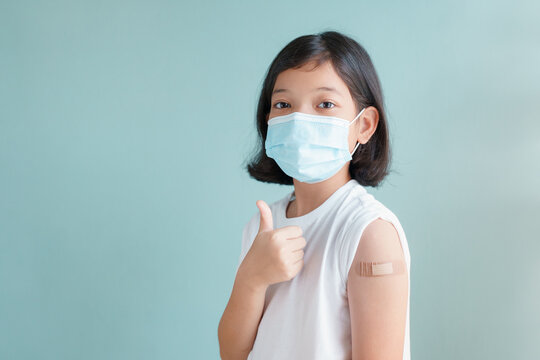 Asian little girl wearing face mask vaccinated Gesturing Thumbs Up  Showing arm bandage to protect COVID-19 spread on blue background