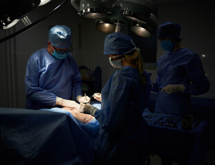 Medical team doing aesthetic plastic surgery in operating room with dim light. Surgeon and...
