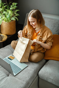 Online Shopping, ordering delivery. Teenager girl relax on sofa considering purchases with laptop. Happy young woman do unpacking online orders goods or food. Mock up paper bags