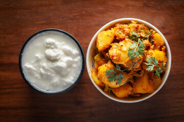 Close-up of Indian vegetarian dish of spicy Potato and Tomato curry garnished with green coriander fresh leaves. Served in a white ceramic bowl over wooden background.
