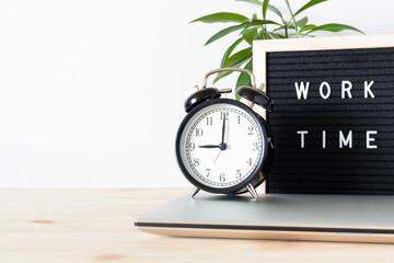 Classic black alarm clock, laptop and black board with words 'work time' on wooden table with white...