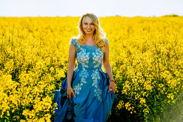 Naklejka na ściany i meble Fashion beautiful young woman blue dress and yellow flowers posing outdoor rapeseed field