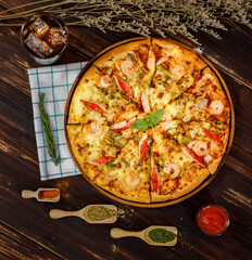 Close up on homemade seafood pizza with shrimp, bell pepper, crab sticks on wooden table with cold beverage, napkin, ingredient of tomato sauce or ketchup, dried plants, paprika chili powder, oregano.