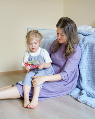 Charming little girl daughter playing with paper educational toy or paper book while sitting on mothers knees and spending leisure time together at home. Parenthood and child development concept