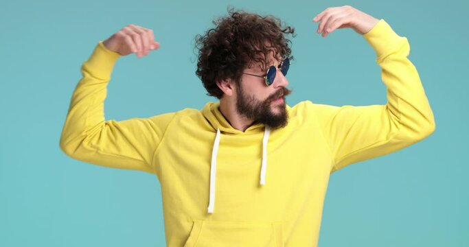 sexy long bearded guy in yellow sweatshirt with retro sunglasses showing muscles and flexing, feeling powerful and smiling on blue background in studio