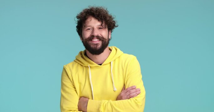 cool long bearded guy in yellow hoodie smiling, nodding and crossing arms while standing on blue background in studio