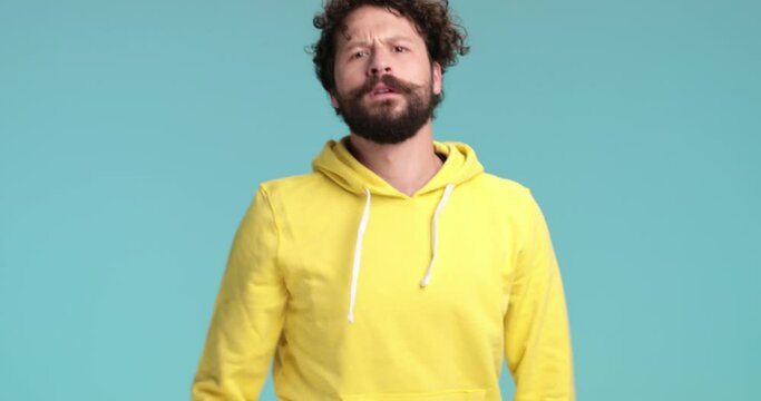 cool young hipster man with long beard and curly hair fighting, holding fists up, pointing fingers and threatening while standing against blue background in studio