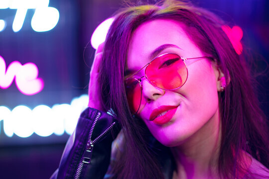 Fashion portrait of a young woman in sunglasses posing near neon signs in night club