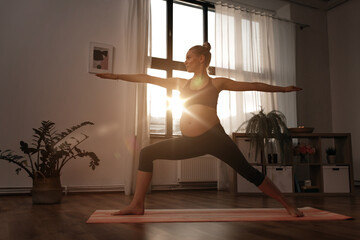 sport, fitness and people concept - happy pregnant woman doing yoga warrior pose at home over sunshine - obrazy, fototapety, plakaty