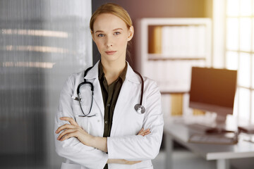 Friendly smiling female doctor standing in sunny clinic. Portrait of friendly physician woman. Medicine concept