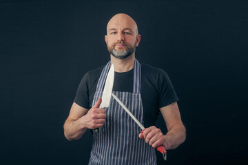 Bald male butcher or fishmonger in black t shirt and classic black and white apron on dark background. Meat industry. Sharpening knife on a metal steel. Man in his 40s, grey beard lean body type