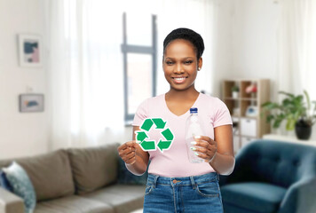 eco living, environment and sustainability concept - smiling young african american woman holding green recycling sign and plastic bottle over home room background