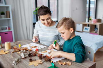 family, creativity and craft concept - mother and little son with glue sticks and paper making pictures of dry autumn leaves, pine cones and chestnuts at home