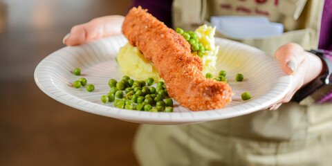 Fish stick with mashed potatoes and green peas served by waiter in restaurant or diner.
