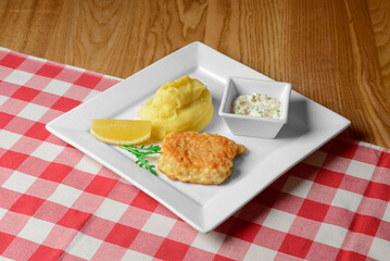 Fried fish with mashed potatoes served in a white square plate over red plaid table cloth.