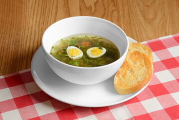 Ukrainian green borsch with boiled eggs served in white plate over red plad table cloth.
