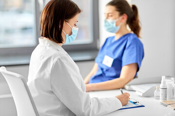 Obraz premium medicine, healthcare and pandemic concept - female doctor in medical protective mask with clipboard and nurse or medical worker at hospital
