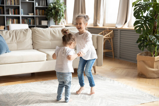 Overjoyed two small preschooler girls children have fun dancing together in cozy living room holding hands. Smiling little sisters kids jump listen to music play at home on weekend. Family concept.