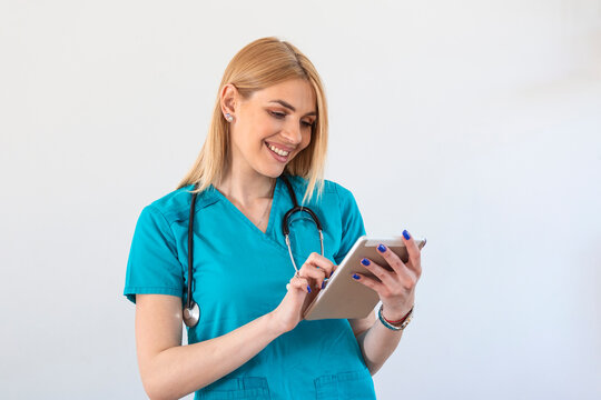 Young female clinician doctor in scrubs using touchpad while communicating with patients online. Portrait Of Smiling Female Doctor Wearing Scrubs In Hospital Corridor Holding Digital Tablet