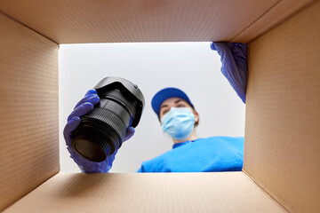 delivery, shipping and pandemic concept - woman in protective medical mask and gloves packing digital camera and lens into parcel box with foam peanuts