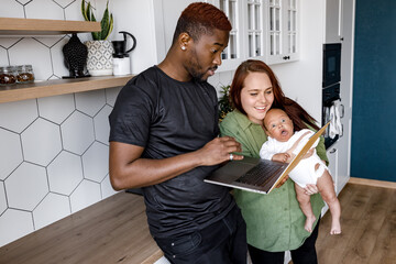 Smiling multiethnic parents relax at home with cute little biracial son using laptop together, happy international family with small boy child stay in kitchen watch video or talk with family online