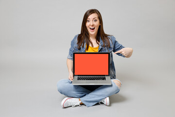 Full length young woman in denim jacket yellow t-shirt point index finger on laptop pc computer blank screen workspace area browsing sitting cross-legged isolated on grey background studio portrait