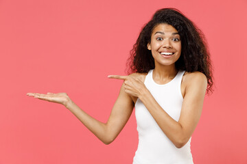 Young fun smiling happy cheerful friendly african american woman 20s wear casual white tank shirt pointing index finger on empty palm with copy space place mock up isolated on pink color background