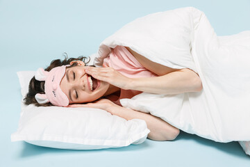 Obraz na płótnie Canvas Young calm woman in pajamas jam sleep eye mask rest relaxing at home lying lies wrap covered under blanket duvet on pillow yawning isolated on pastel blue background. Good mood night bedtime concept.