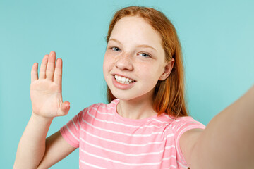 Fototapeta premium Close up little redhead kid girl 12-13 years old in pink striped t-shirt doing selfie shot on mobile phone waving hand hello isolated on pastel blue background. Children lifestyle childhood concept