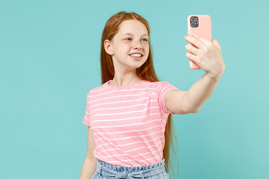 Little redhead kid girl 12-13 years old wearing pink striped tshirt do selfie shot on mobile phone post photo on social network isolated on pastel blue background Children lifestyle childhood concept
