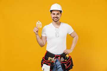 Young smiling employee handyman man 20s in protective helmet hardhat holding house mock up stand akimbo isolated on yellow background Instruments accessories renovation apartment room Repair concept.