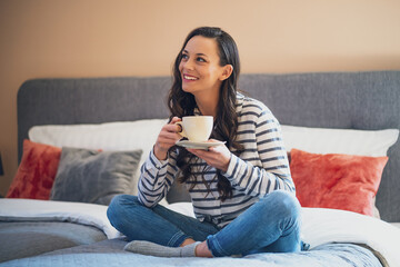 Happy young woman sitting on bed in her home and drinking coffee.