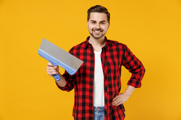 Young smiling employee handyman man in checkered shirt hold putty knife stand akimbo isolated on yellow background studio. Instruments accessories for renovation apartment room. Repair home concept.